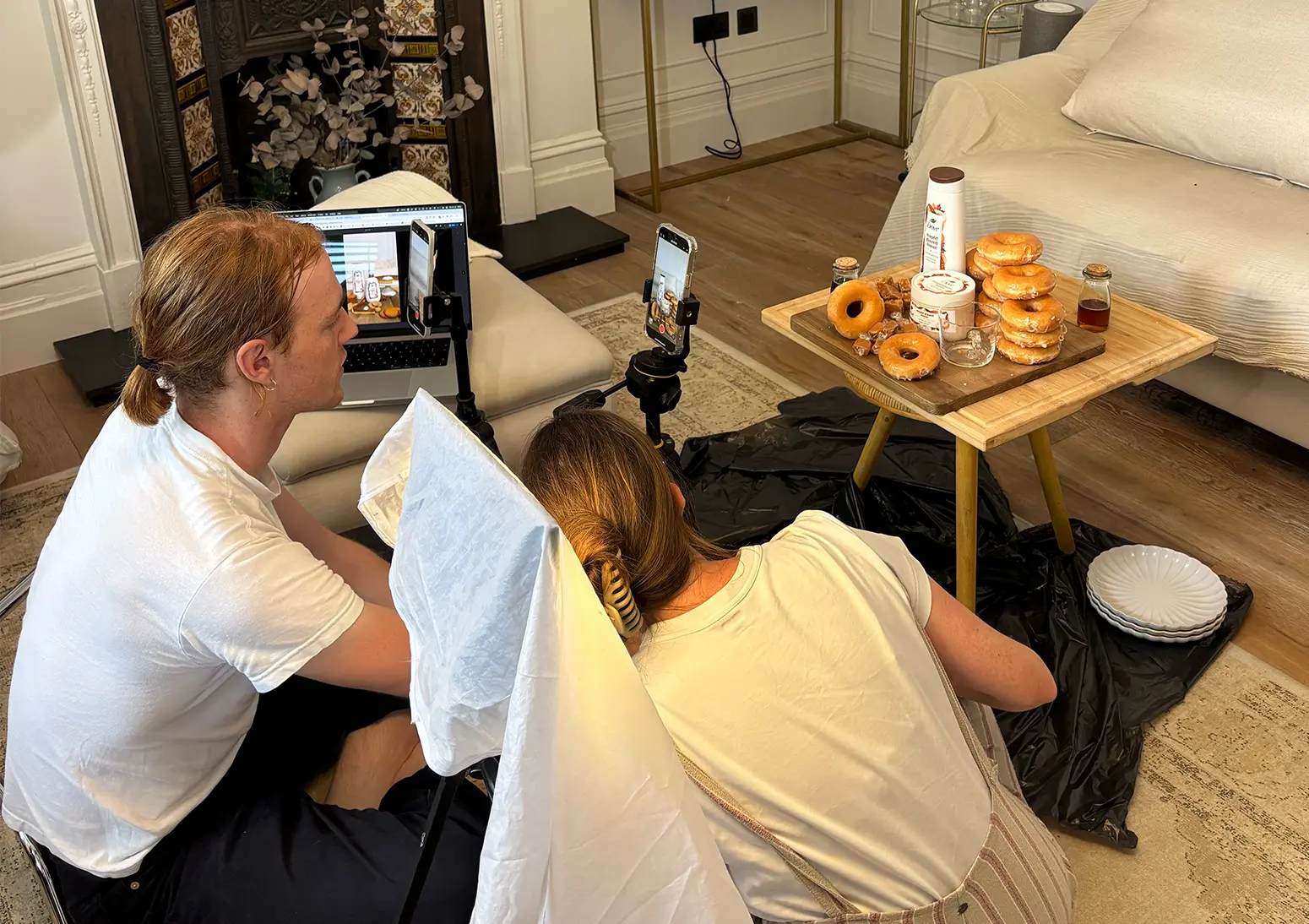 Two people filming a food setup in a living room using smartphones on tripods, with donuts and packaged products arranged on a small table, illustrating a location production setup.